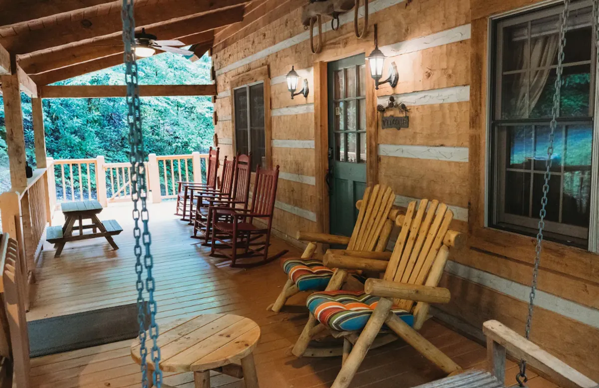 A covered porch with classic rocking chairs, a wooden table, and warm hanging lights, set against the backdrop of surrounding greenery. The scene suggests peaceful relaxation and a strong connection to the outdoors.