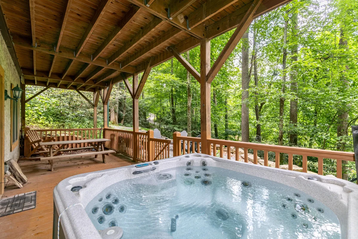 A tranquil outdoor scene featuring a wooden deck nestled among trees, with a hot tub as the focal point. The hot tub is positioned for scenic views and privacy, with ample space for relaxing and socializing. The surrounding greenery enhances the secluded, nature-immersed atmosphere.