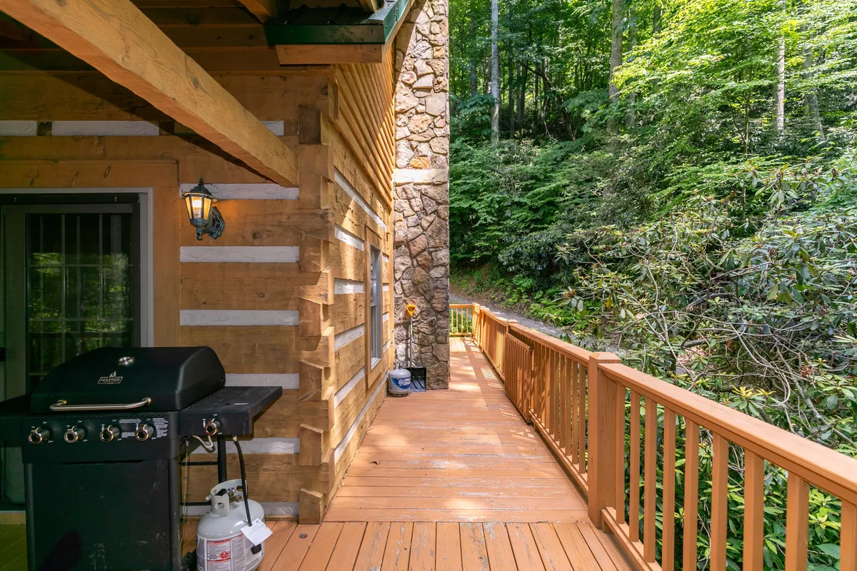 A welcoming wooden deck with a grill and stone wall overlooks vibrant greenery. The inviting outdoor space is ideal for barbecues, enjoying morning coffee, or unwinding after a day of hiking, fishing, or biking.