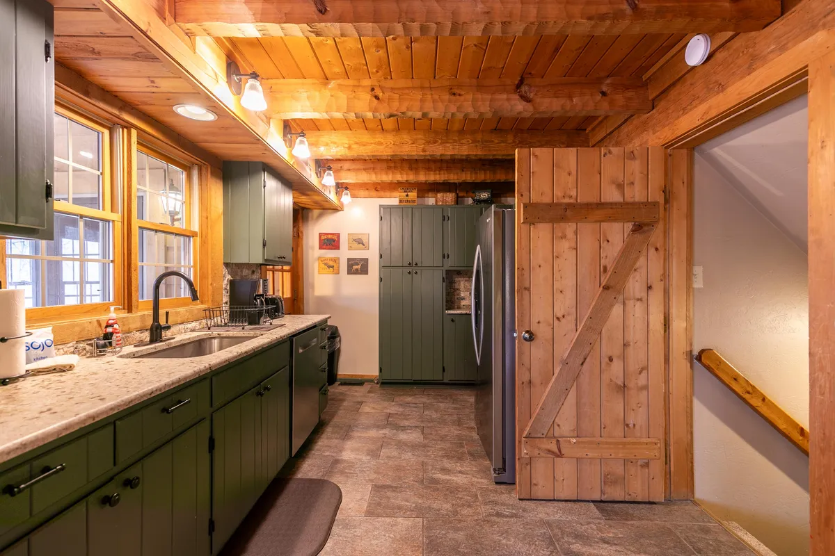 A modern kitchen with a spacious island and striking green cabinetry, complemented by wooden beams and expansive windows. The setup encourages social cooking and flow into adjacent living spaces, embodying the 'fully stocked kitchen' ideal for culinary adventures.
