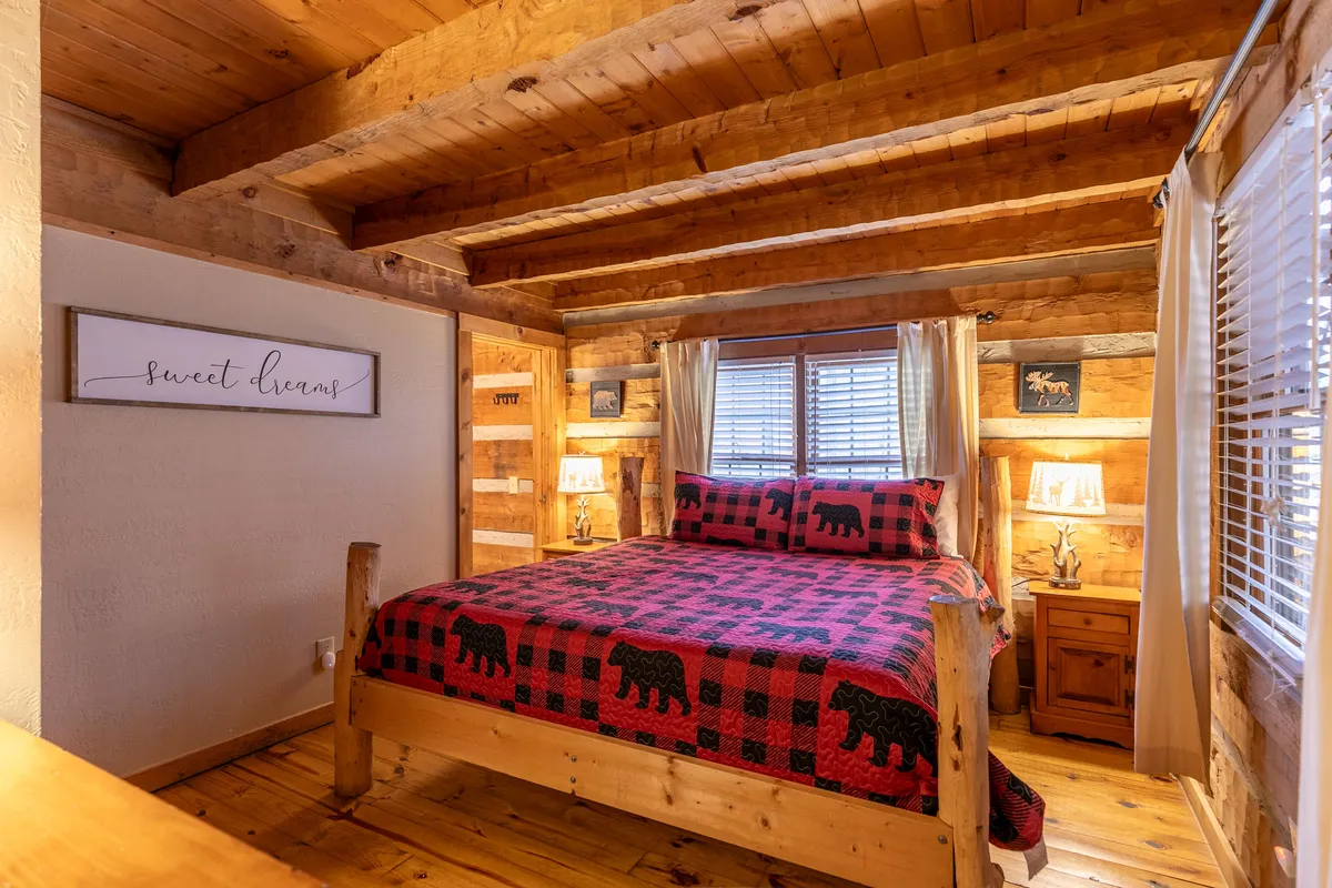 A charming bedroom with bear-patterned bedding on a queen-sized bed, wooden beams overhead, and a window draped with curtains. The room feels intimate and warm, ideal for reading or unwinding.
