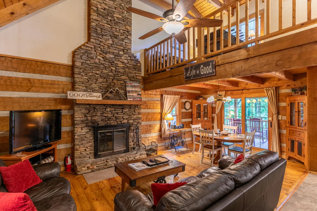 A view into the heart of the cabin’s main level, featuring a substantial stone fireplace surrounded by cozy seating, exposed wooden beams, a welcoming dining area, and large windows that invite natural light. The open layout visually connects the living and dining spaces, creating an atmosphere perfect for gatherings and relaxation.