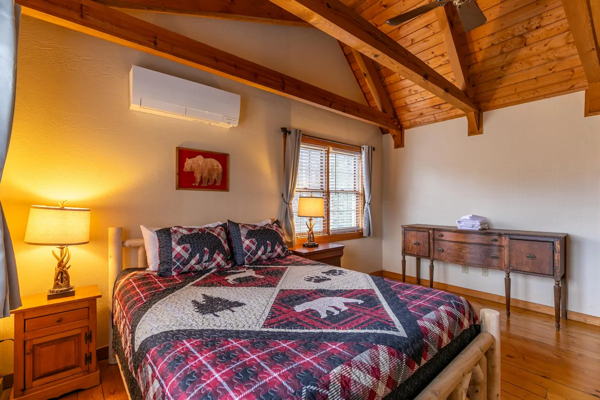 A cozy bedroom featuring a king-sized bed with a patterned quilt, exposed wooden beams, a window with soft curtains, a nightstand with a glowing lamp, and a sturdy dresser. Sunlight streams in, creating a warm and inviting atmosphere ideal for relaxation.