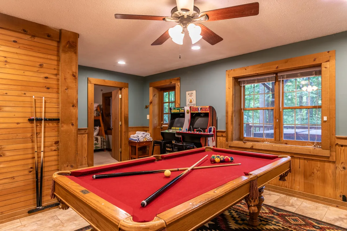 A dedicated game room featuring a pool table, arcade machine, wood paneling, and windows for natural light. The setting is playful and relaxed, offering a perfect environment for friendly competitions and hours of entertainment.