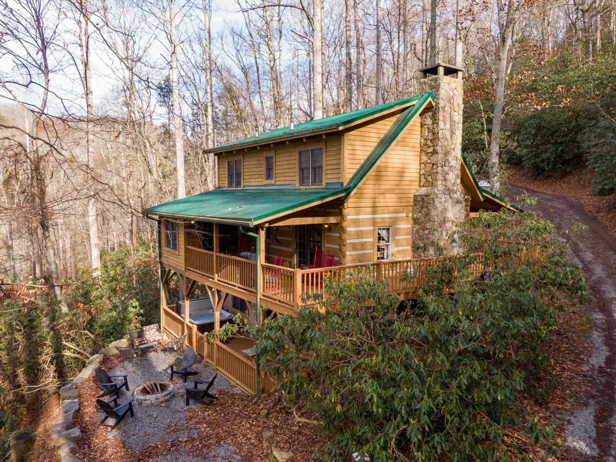 The image showcases a classic log cabin nestled among tall trees, with a sturdy stone chimney and a wraparound deck framed by rustic railings. A cozy fire pit sits in the foreground, inviting guests to gather under the stars, while the wooded surroundings hint at peaceful seclusion and nature-filled adventures.
