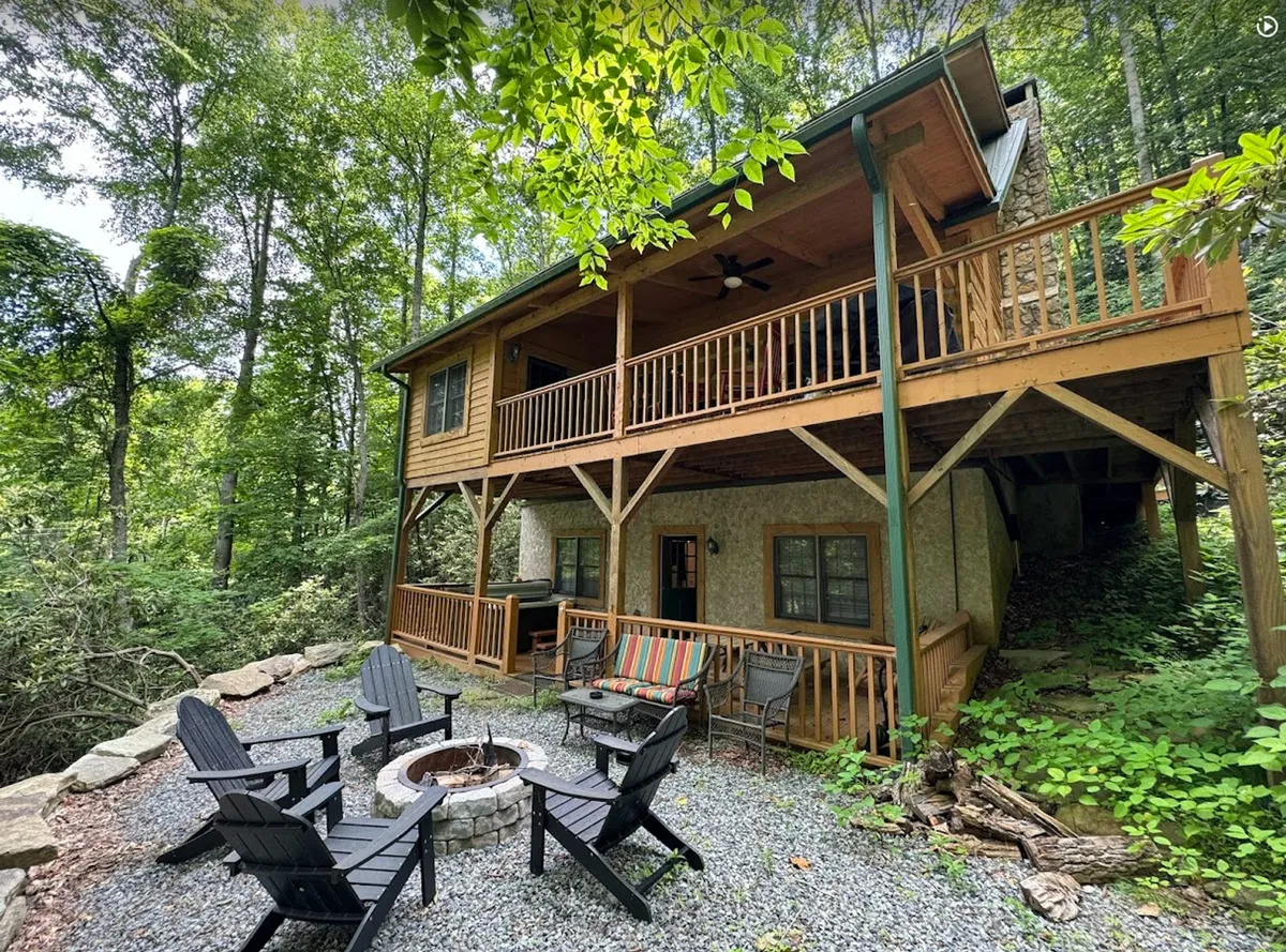 A wide exterior view of the cabin with a fire pit, wooden deck, and seating area, all surrounded by dense trees. The scene exudes seclusion, comfort, and connection with nature, illustrating the overall setting and the opportunities for group gatherings.