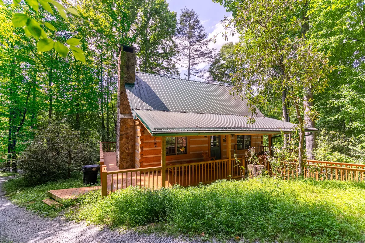 A classic log cabin nestled in vibrant green foliage, featuring a spacious wooden deck that blends seamlessly into its natural woodland setting. The cabin's rustic charm is highlighted by the warm wood tones and the surrounding trees, suggesting ample space for outdoor activities and walks.