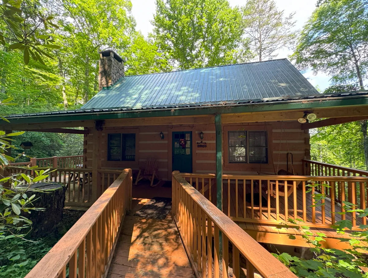 The log cabin’s rustic exterior features a sturdy stone chimney and a charming porch set alongside a quaint wooden bridge. The greenery and natural surroundings evoke a sense of seclusion and adventure, with the porch offering prime wildlife viewing opportunities.
