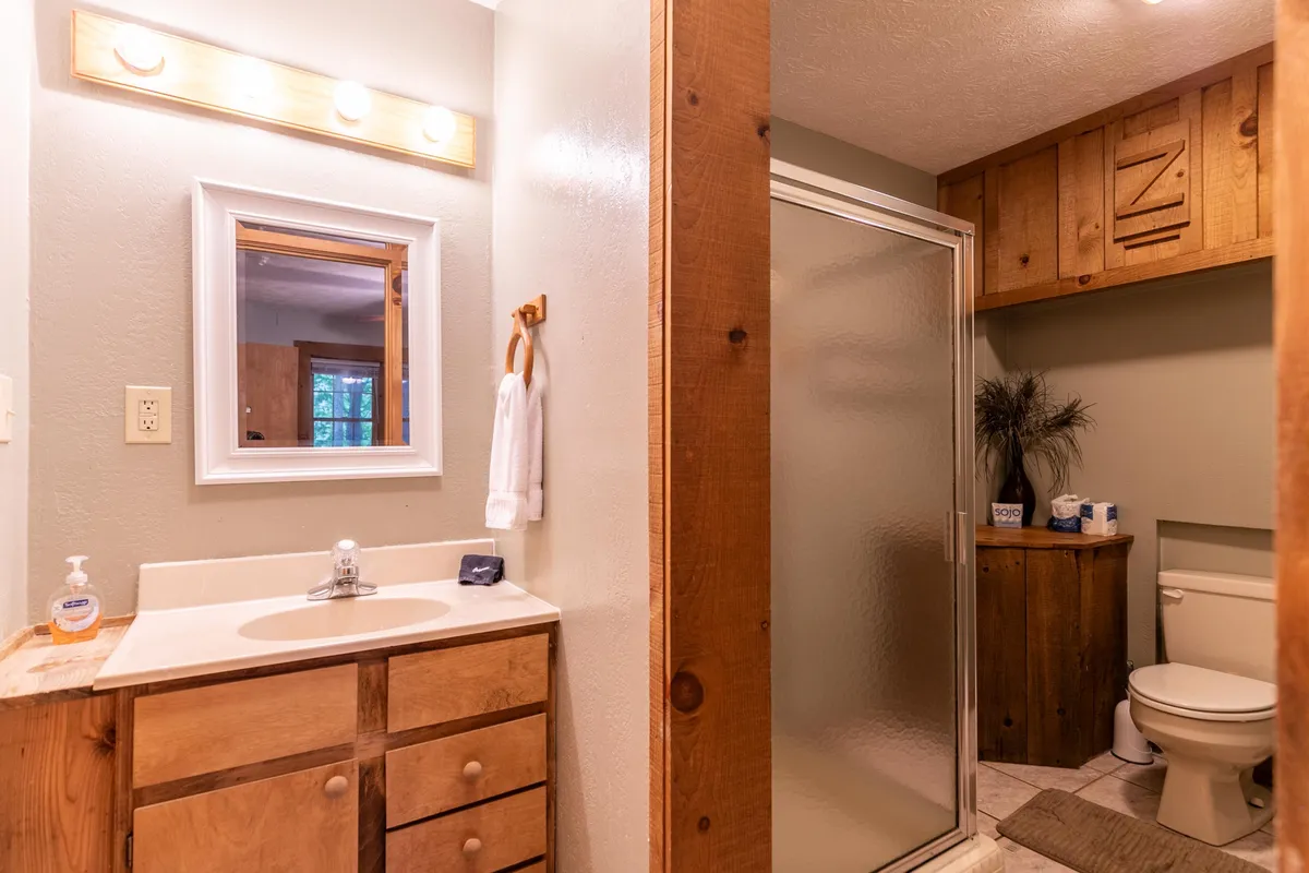 A practical, well-appointed bathroom with a vanity, wooden cabinets, mirror, shower enclosure, and toilet area. Sunlight streams in, showcasing a clean and inviting space for freshening up after the day’s activities.