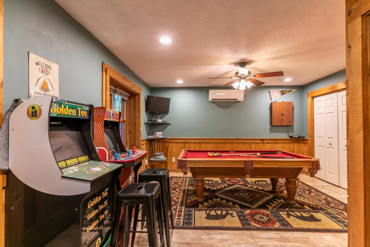 This image features a lively game room with a pool table at the center, flanked by two colorful arcade machines and a cozy decorative rug beneath. The warm lighting and casual atmosphere create an inviting environment designed for fun and friendly competition.