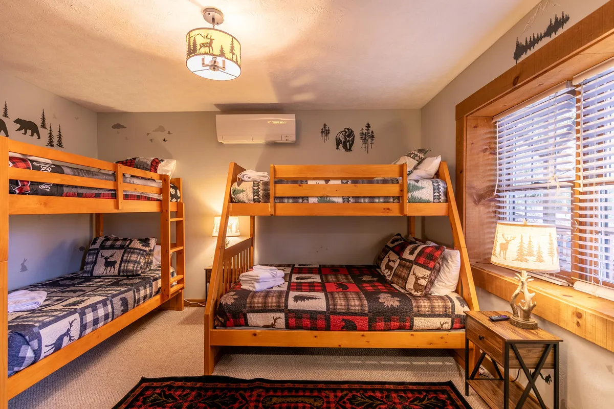 A cozy bunk room is filled with multiple beds featuring textured blankets and decorative wildlife lamps. The playful wall decals and warm lighting make it inviting for kids and families alike—a sleepover-ready space with character.